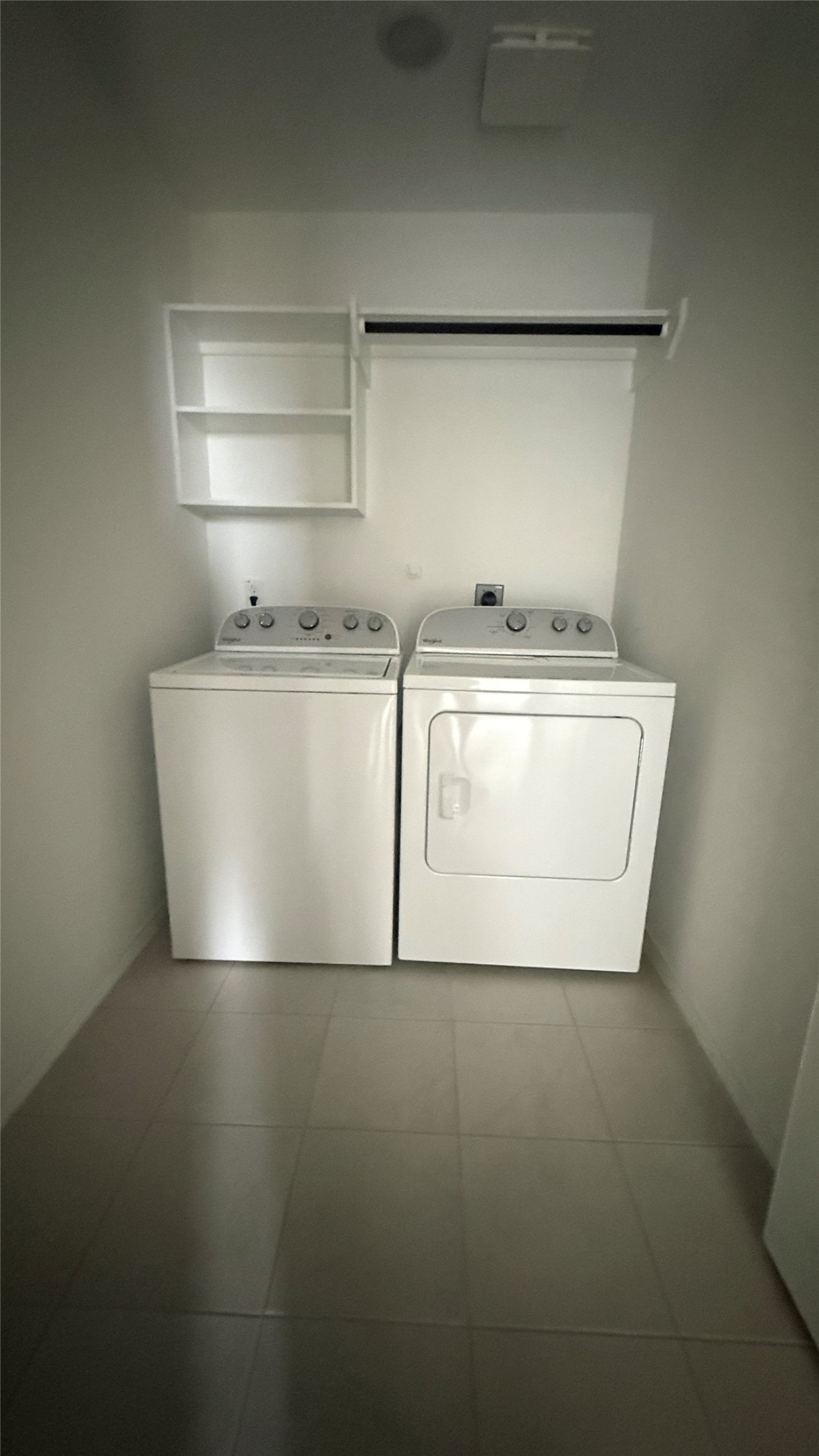 31961 Retama Rnch Drive Spring, TX 77386 - Photo 6 of 25 This photo shows a compact laundry room with a washer and dryer side by side. It features tiled flooring, a wall-mounted shelf, and a hanging rod for convenient storage.