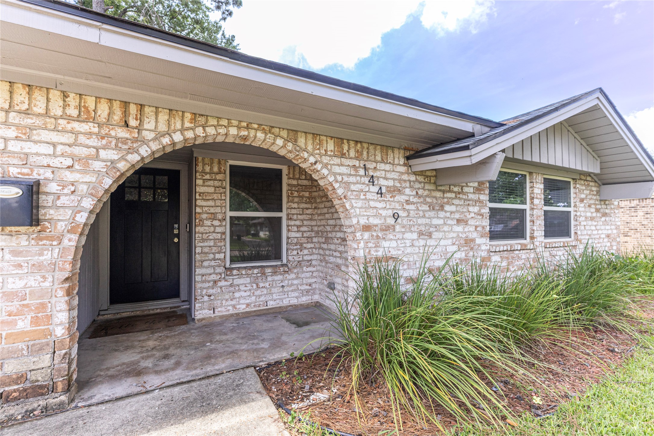14419 Wadebridge Way Houston, TX 77015 - Photo 18 of 20 This photo features a charming brick home with a welcoming arched entryway and a modern black front door. The exterior is accented by well-kept landscaping with green shrubs lining the walkway. The home has a classic, inviting appearance.