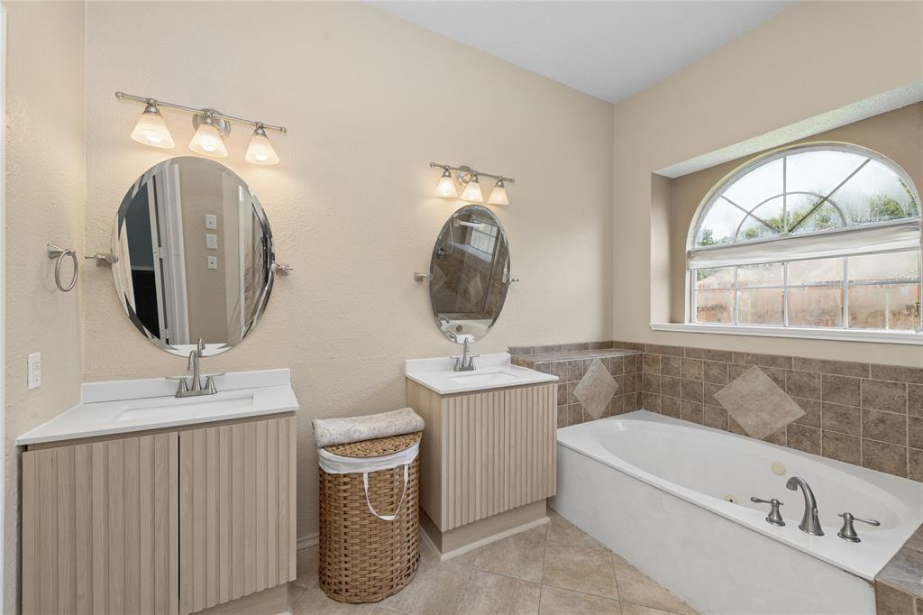 4202 Timberglen Road Dallas, TX 75287 - Photo 23 of 40 Bathroom featuring a built-in soaking tub, arched window, and dual vanity areas with oval mirrors and light-toned cabinetry