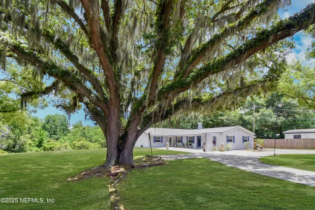 a house that is sitting in the grass with large trees and plants