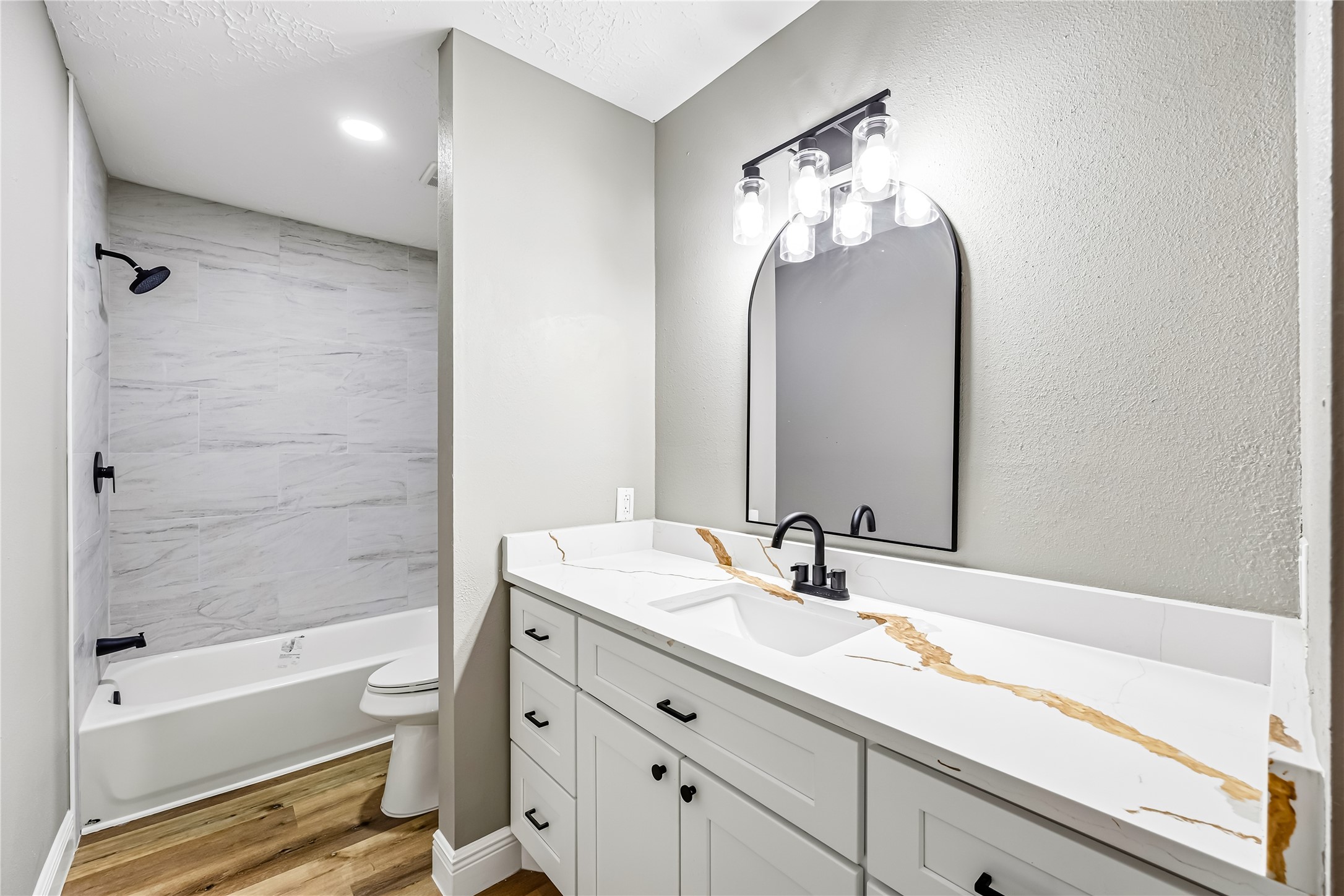 4534 Rosegate Drive Spring, TX 77373 - Photo 14 of 24 Updated bathroom featuring a modern vanity, striking stone-look countertop, black fixtures, arched mirror, and contemporary lighting. The tiled tub and shower surround, neutral finishes, and warm wood-look flooring create a clean, stylish, and inviting space.