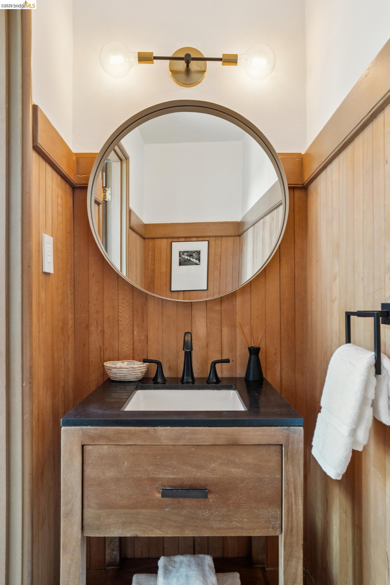 38 Highgate Road Kensington, CA 94707 - Photo 11 of 78 Wood paneling, a circular mirror, a wood-finish vanity, a dark countertop, and a matte black faucet