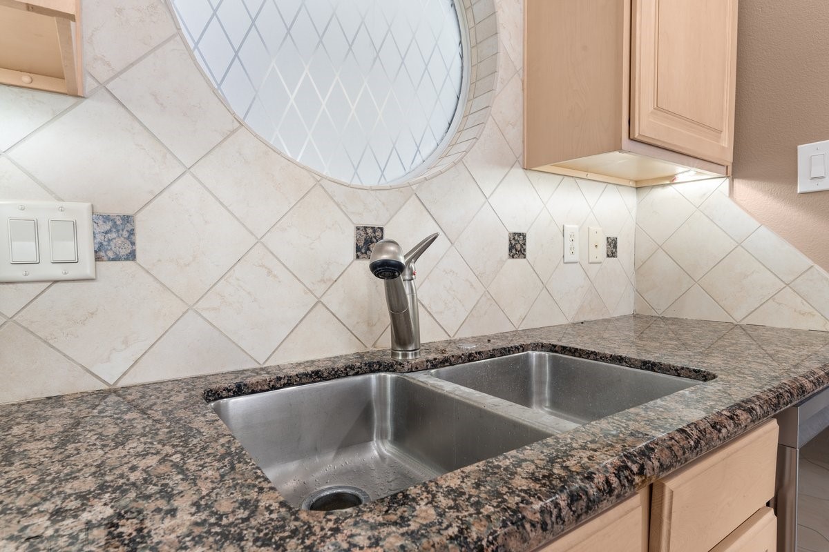 168 Lake Point Boulevard, Unit C102 Conroe, TX 77356 - Photo 8 of 35 This kitchen features a double stainless steel sink set in a granite countertop, with light wood cabinets and a decorative tile backsplash, including a unique round window for added character.