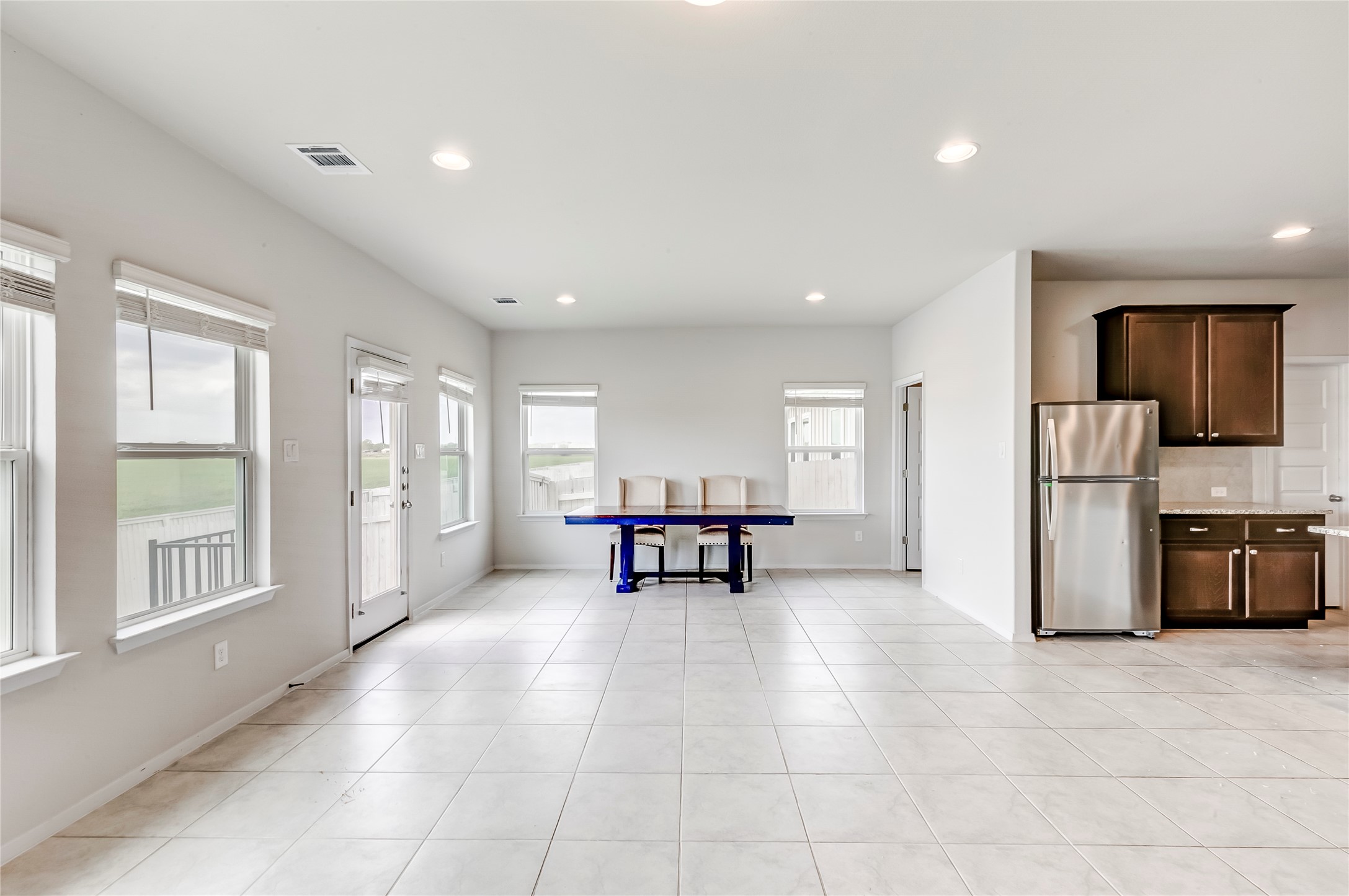 2721 Winding Creek Road Kyle, TX 78640 - Photo 7 of 27 Spacious open-concept room featuring light-toned tile flooring, recessed lighting, multiple windows with white blinds, and a kitchen area with dark wood cabinetry and a stainless steel refrigerator