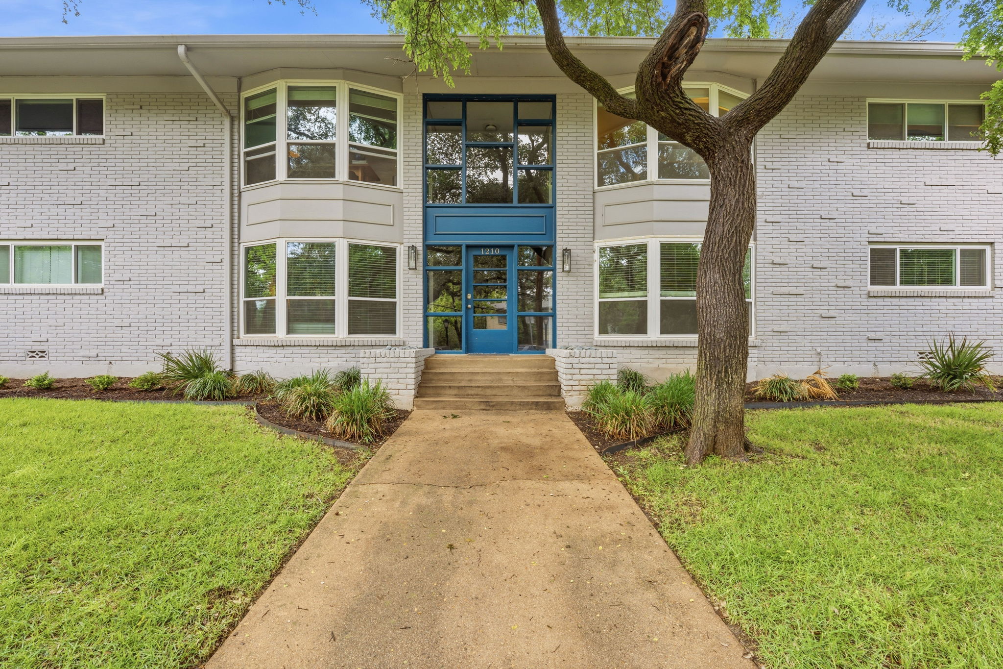 1210 Windsor Road, Unit 104 Austin, TX 78703 - Photo 2 of 27 Brick facade building with a prominent blue entrance, featuring a concrete pathway and planted landscaping