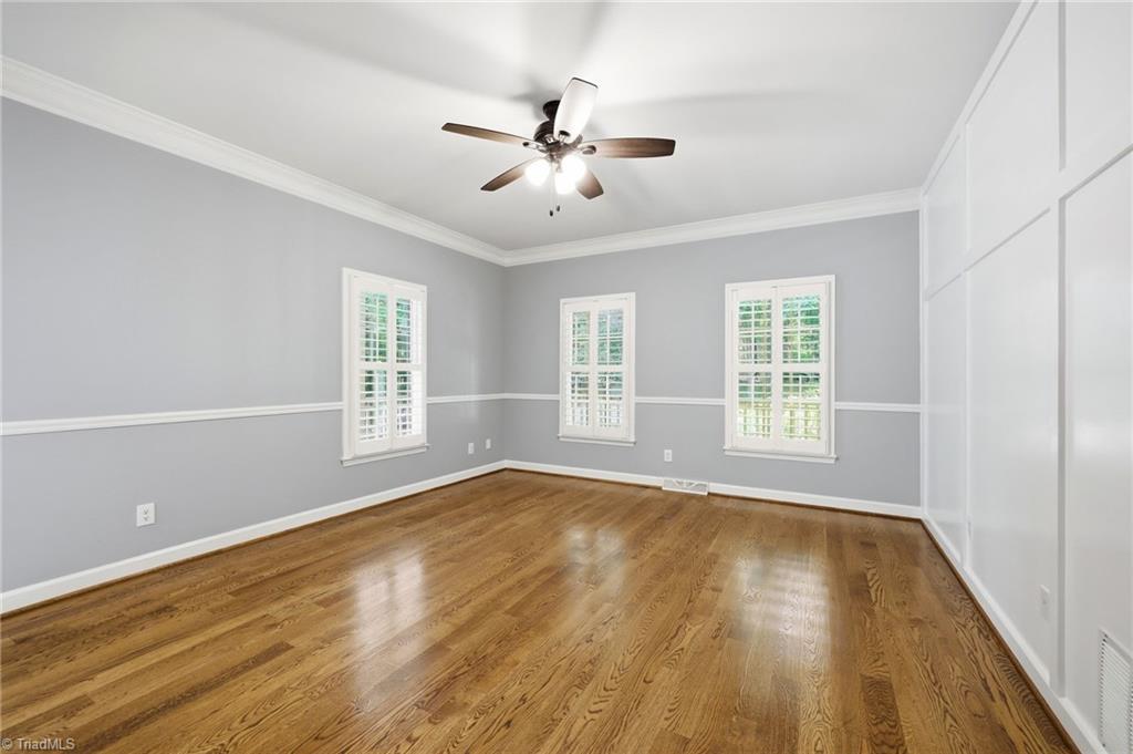 8861 Boylston Road Colfax, NC 27235 - Photo 18 of 50 The spacious main-level primary suite offers a peaceful retreat with rich hardwood floors, elegant board-and-batten accent wall, crown molding, and abundant natural light. The generous layout easily accommodates large furnishings while providing direct access to the ensuite bath, creating a comfortable and private owner's retreat.