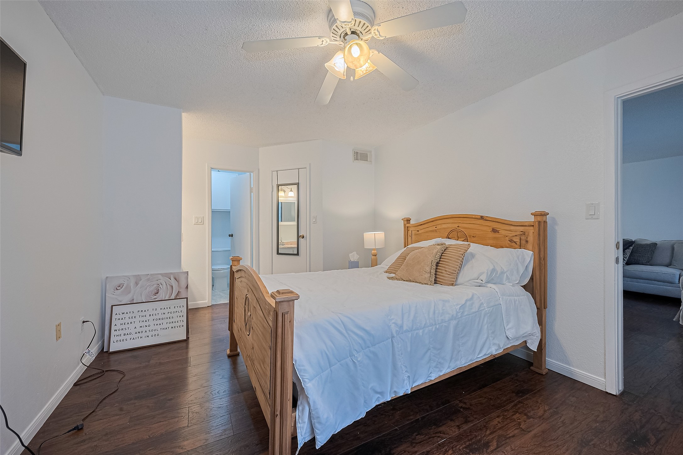 1505 Ward Road, Unit 118 Baytown, TX 77520 - Photo 19 of 35 Cozy bedroom with wood flooring, featuring a ceiling fan and neutral walls. It includes a wooden dresser, wall-mounted TV, and a bed with plush pillows. An en-suite bathroom and closet are accessible through an open door.