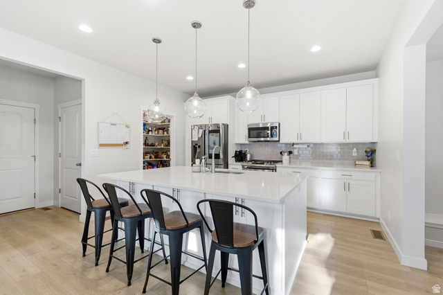 1038 South Old Towne Road Layton, UT 84041 - Photo 6 of 39 Modern kitchen featuring a large central island with seating for four, white cabinetry, integrated stainless steel appliances, a subway tile backsplash, and light wood-finish flooring