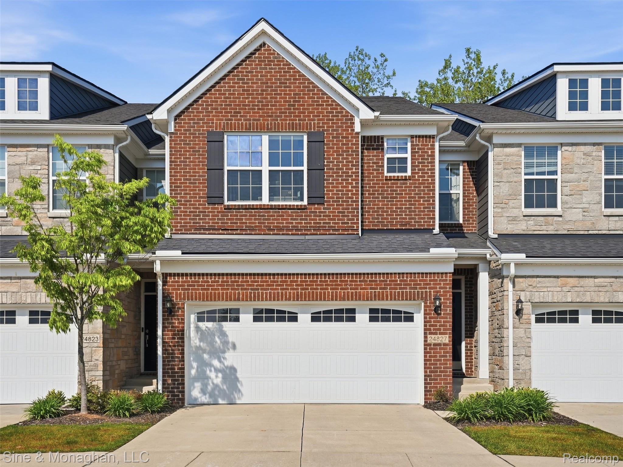 24827 Ivy Road Novi, MI 48375 - Photo 1 of 38 Brick and stone facade townhome featuring a two-car garage with arched windows, black decorative shutters, and gabled dormers