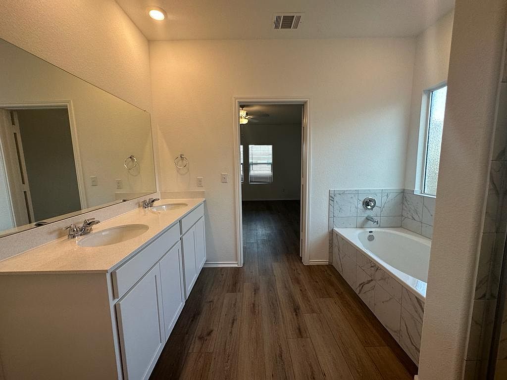 160 River Rise Road Kyle, TX 78640 - Photo 14 of 35 Contemporary bathroom featuring a double vanity with ample counter space, a large mirror, a built-in tub with marble-look tile surround, and wood-finish flooring