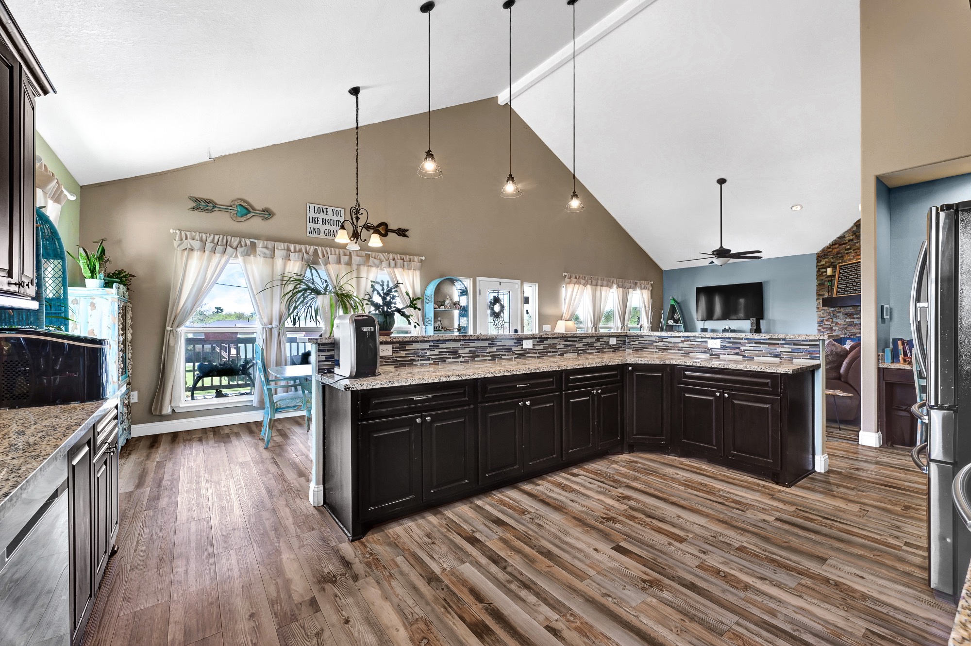 939 8th Street Dickinson, TX 77539 - Photo 11 of 31 This photo showcases a spacious, open-plan kitchen with high ceilings and ample natural light. It features dark wood cabinetry, modern pendant lighting, and a large countertop with a stylish tiled backsplash. The kitchen opens to a cozy living area, creating an inviting space for entertaining.
