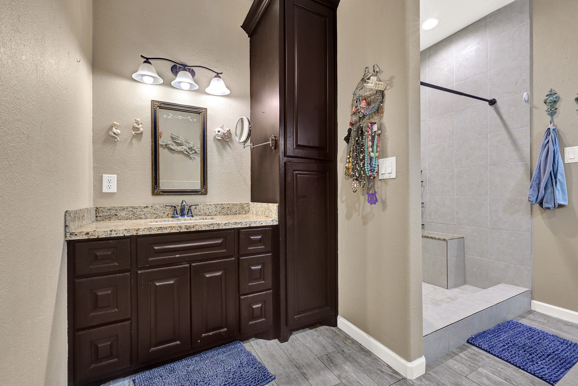 939 8th Street Dickinson, TX 77539 - Photo 23 of 31 This bathroom features dark wood cabinetry with a granite countertop and a framed mirror. It has a spacious walk-in shower with neutral tile. The space is well-lit with a three-light fixture above the sink.