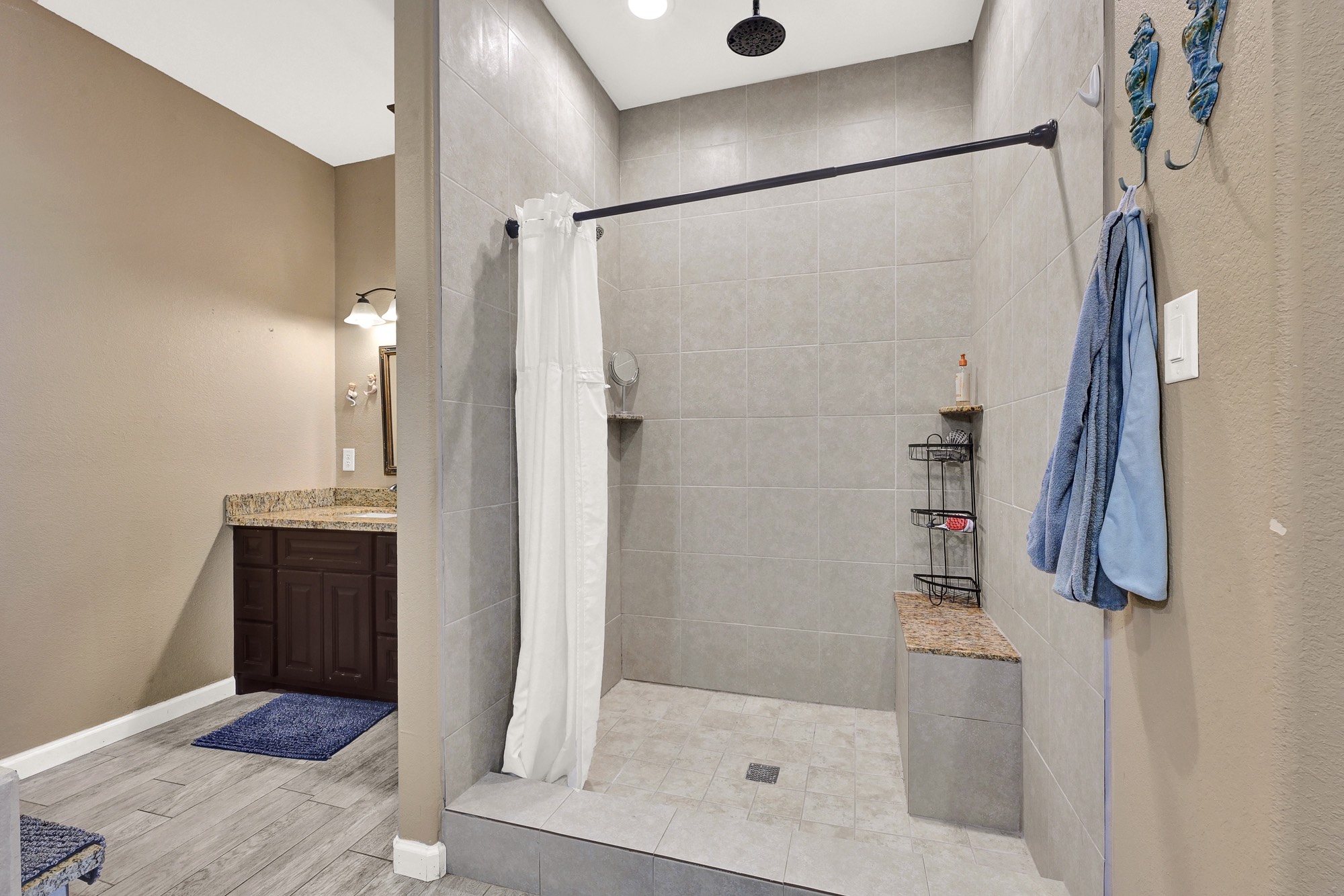 939 8th Street Dickinson, TX 77539 - Photo 24 of 31 Modern bathroom featuring a spacious, open walk-in shower with neutral tile and a rainfall showerhead. There’s a granite bench and shelving for storage. The vanity area includes a granite countertop and dark wood cabinetry, complemented by soft lighting and a cozy color scheme.
