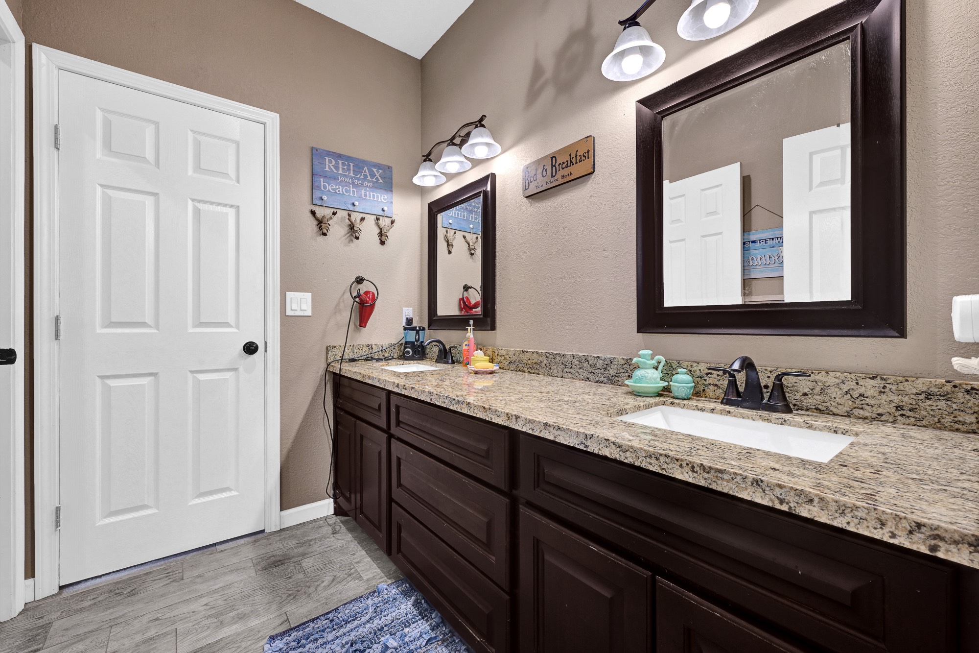 939 8th Street Dickinson, TX 77539 - Photo 28 of 31 This bathroom features a double vanity with granite countertops, dark wood cabinetry, and stylish light fixtures. The neutral wall color complements the wood-look tile flooring, creating a cozy and inviting atmosphere.
