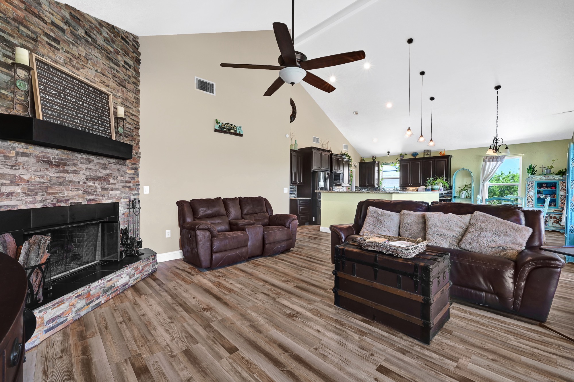 939 8th Street Dickinson, TX 77539 - Photo 10 of 31 This spacious living area features a high vaulted ceiling with a ceiling fan, a cozy stone fireplace, and wood flooring. The open-concept design seamlessly connects to a modern kitchen with dark cabinetry and stylish lighting, creating a warm and inviting atmosphere.