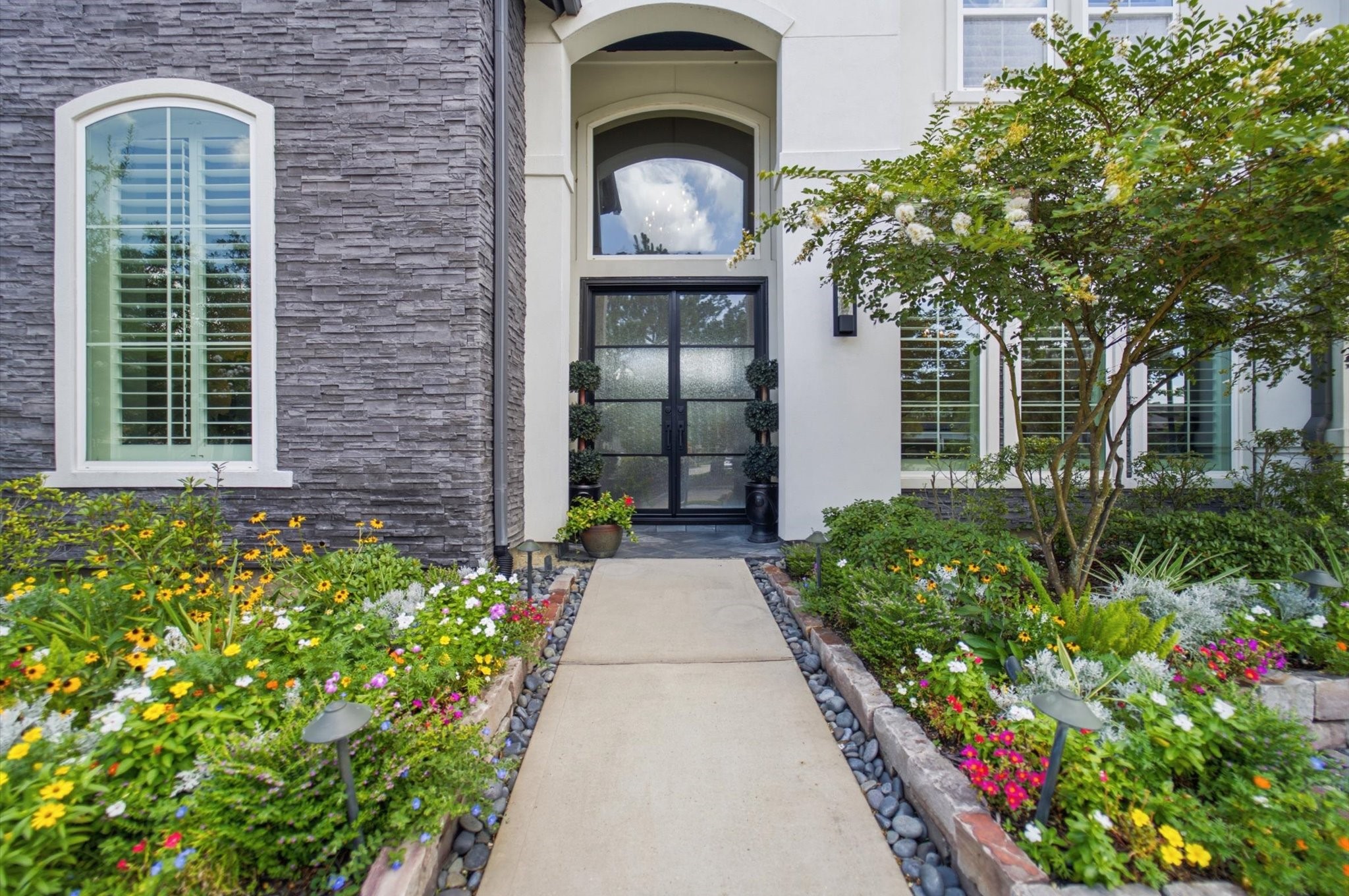 23722 Thortons Park Lane Spring, TX 77389 - Photo 4 of 48 This welcoming entrance features a modern black wrought iron door framed by elegant stone and stucco, surrounded by vibrant, well-maintained flower beds and a clean walkway.