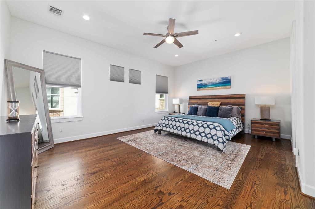 710 Delano Street, Unit A Houston, TX 77003 - Photo 7 of 32 This spacious primary bedroom features hardwood floors, a large bed with modern bedding, and a ceiling fan. It includes a stylish dresser, a large mirror, and ample natural light from multiple windows. The room's neutral tones create a calming atmosphere.
