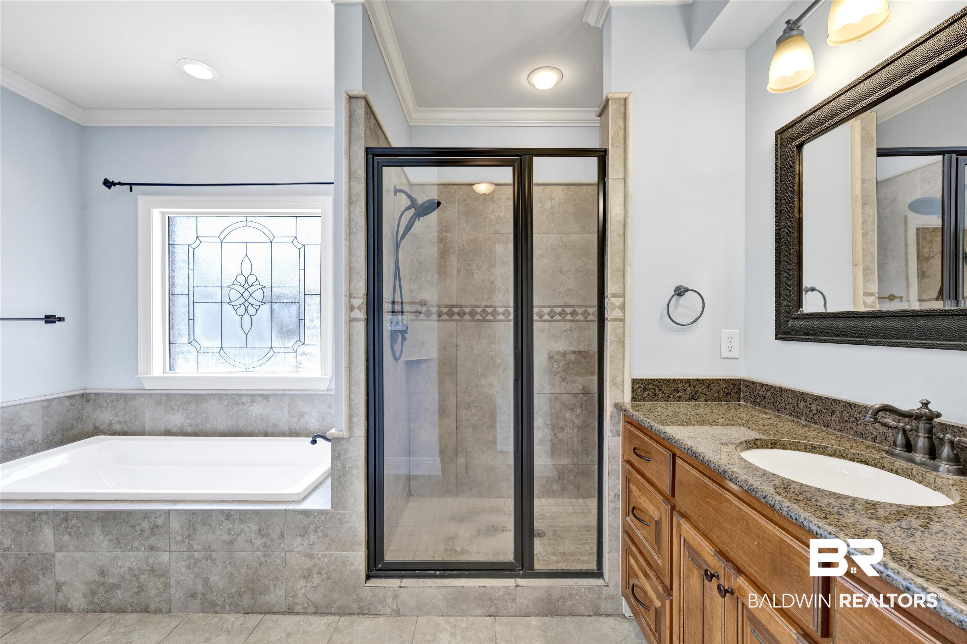 12944 Anna's Way Stapleton, AL 36578 - Photo 50 of 64 Bathroom with a corner soaking tub, a tiled walk-in shower with a glass enclosure, a leaded glass window, and a vanity with a granite countertop and wood-finish cabinetry
