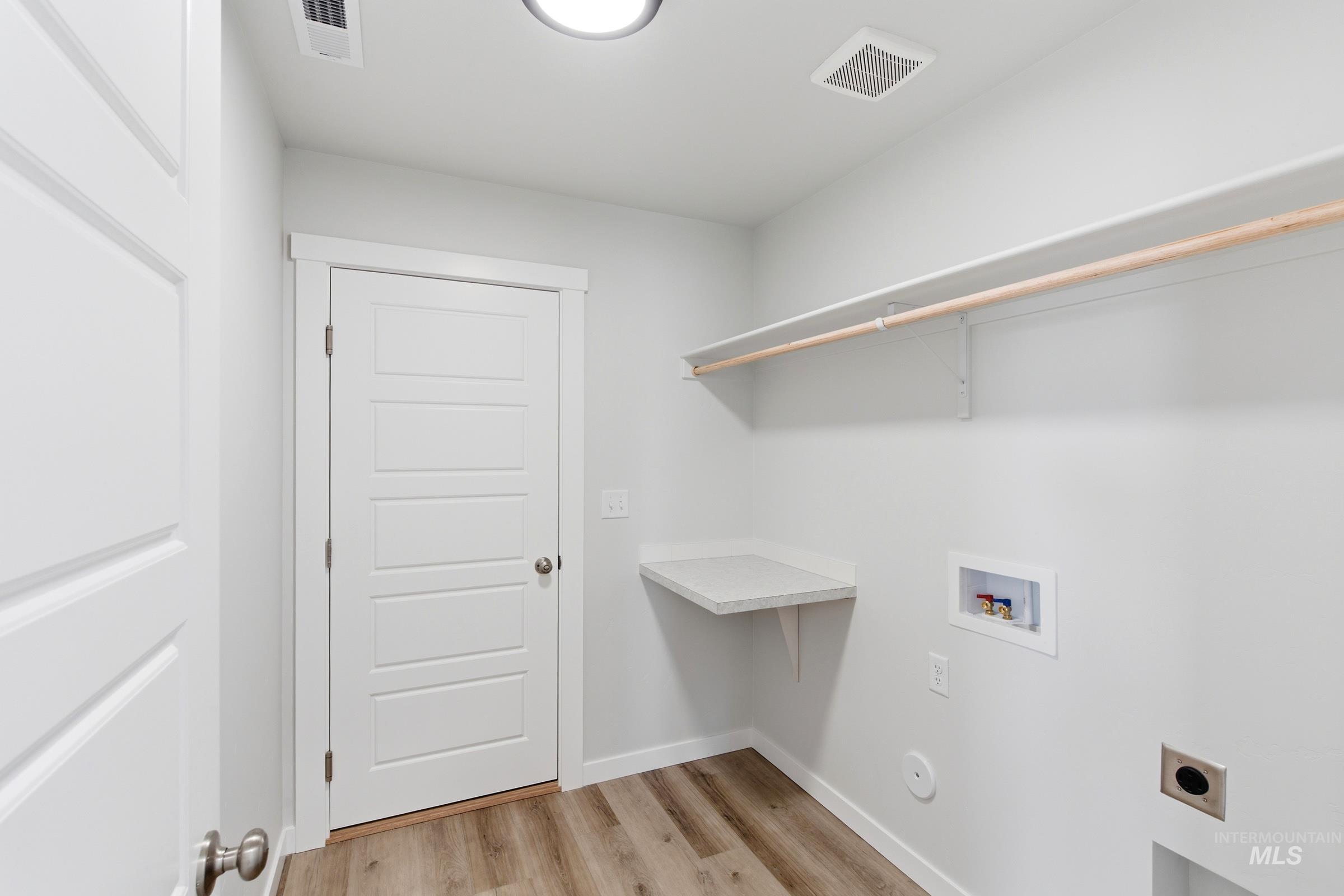8317 South Tyrion Way Kuna, ID 83634 - Photo 9 of 22 Laundry room featuring wood-finish flooring, a white paneled door, a wall-mounted folding counter, and an overhead shelf with a hanging rod
