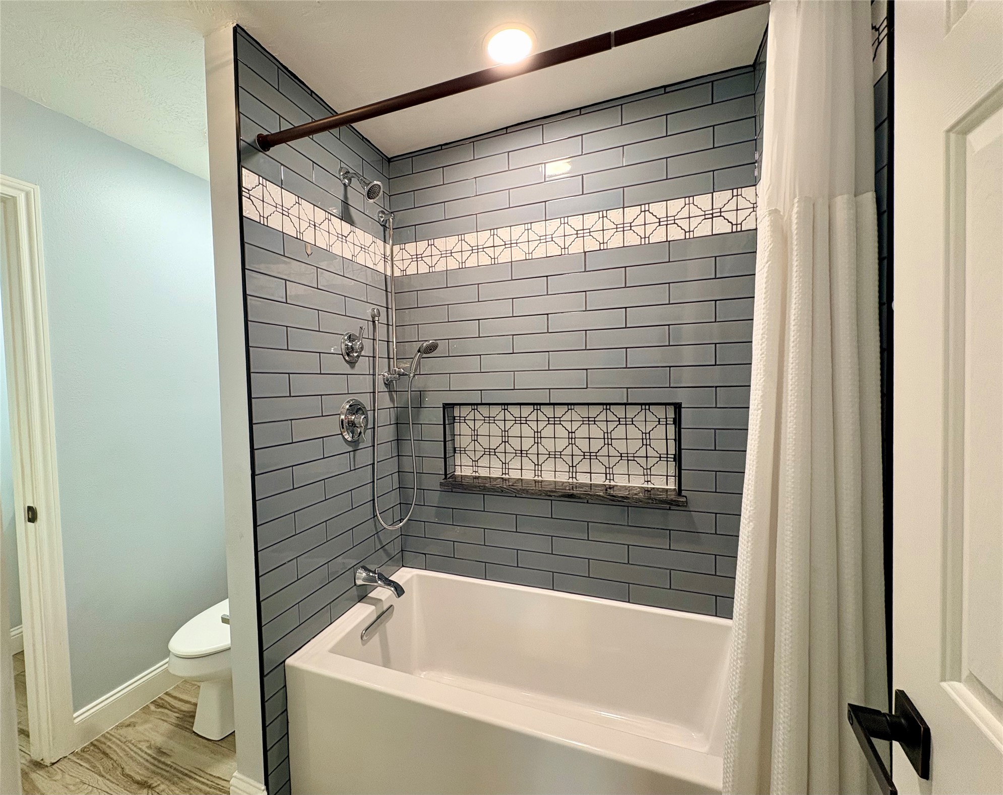 15410 Redbud Berry Way Cypress, TX 77433 - Photo 36 of 49 This bathroom features a modern tub-shower combo with sleek gray subway tiles, accented by a decorative band. It includes a built-in shelf and a curtain rod, set against a light wall and wooden flooring.