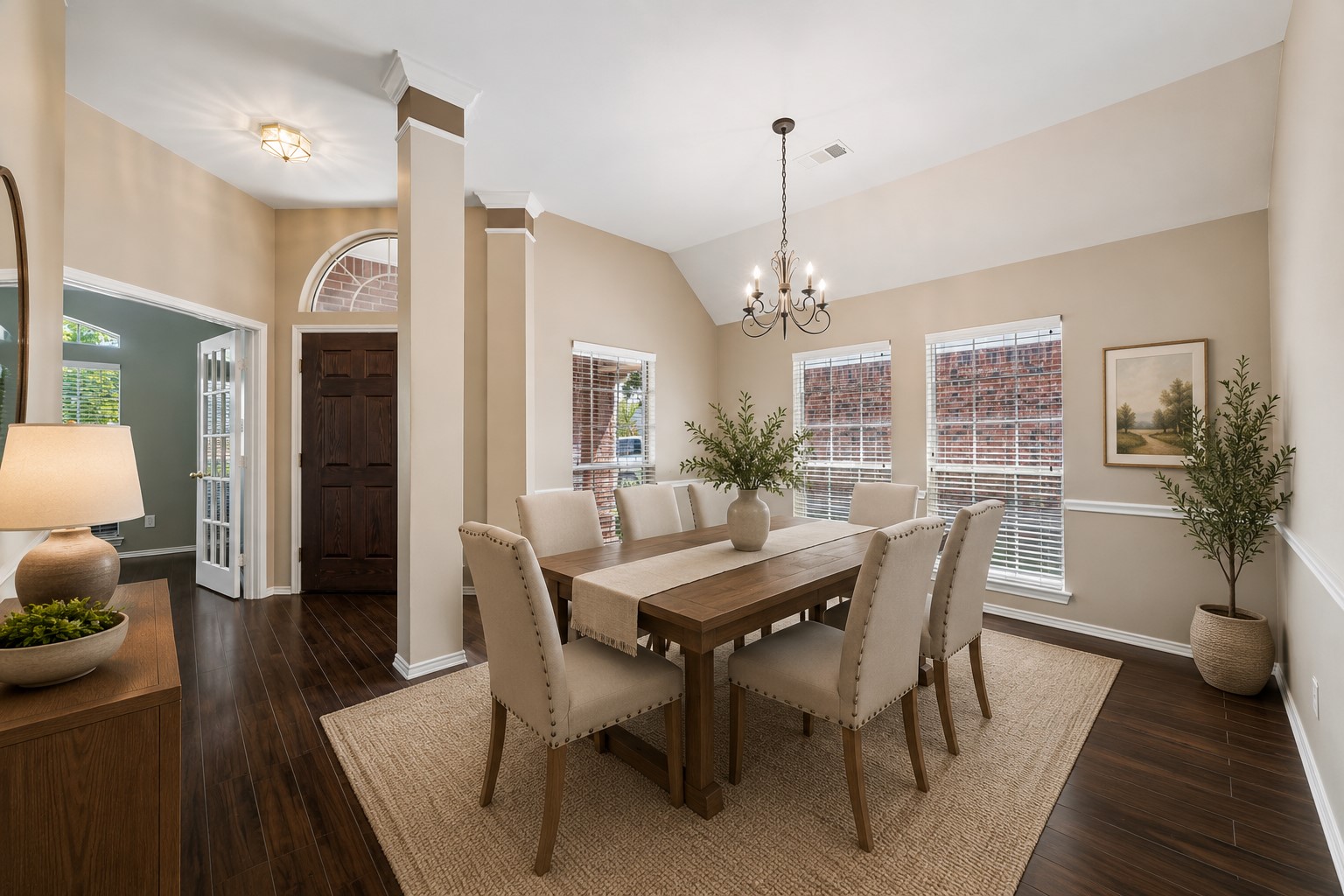 3118 Valley Court Manvel, TX 77578 - Photo 4 of 38 This photo showcases a virtual staged spacious dining room with a vaulted ceiling and elegant chandelier. It features a large wooden table surrounded by cushioned chairs. The room is well-lit with natural light from three windows, complemented by warm, neutral walls and dark hardwood flooring. A decorative plant and framed artwork add charm to the space.