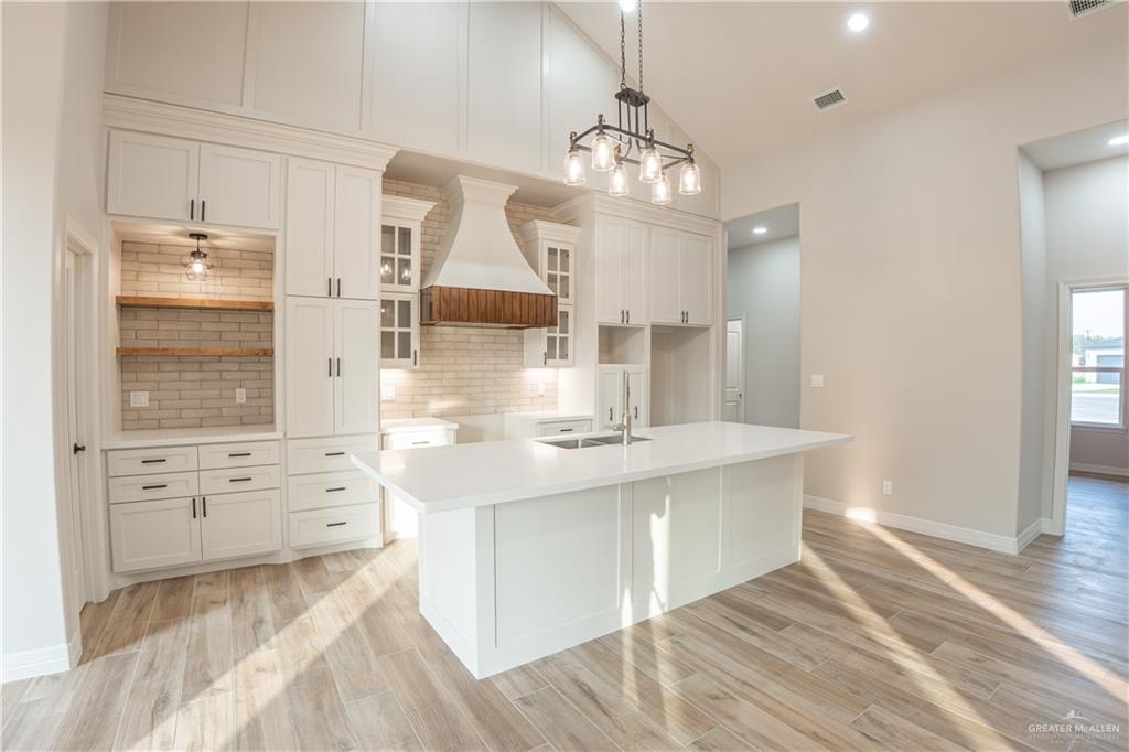 2618 Max Street Harlingen, TX 78552 - Photo 13 of 35 Spacious kitchen featuring white cabinetry, a large center island with a sink, light-toned wood-finish flooring, a decorative range hood with wood accents, and a subway tile backsplash
