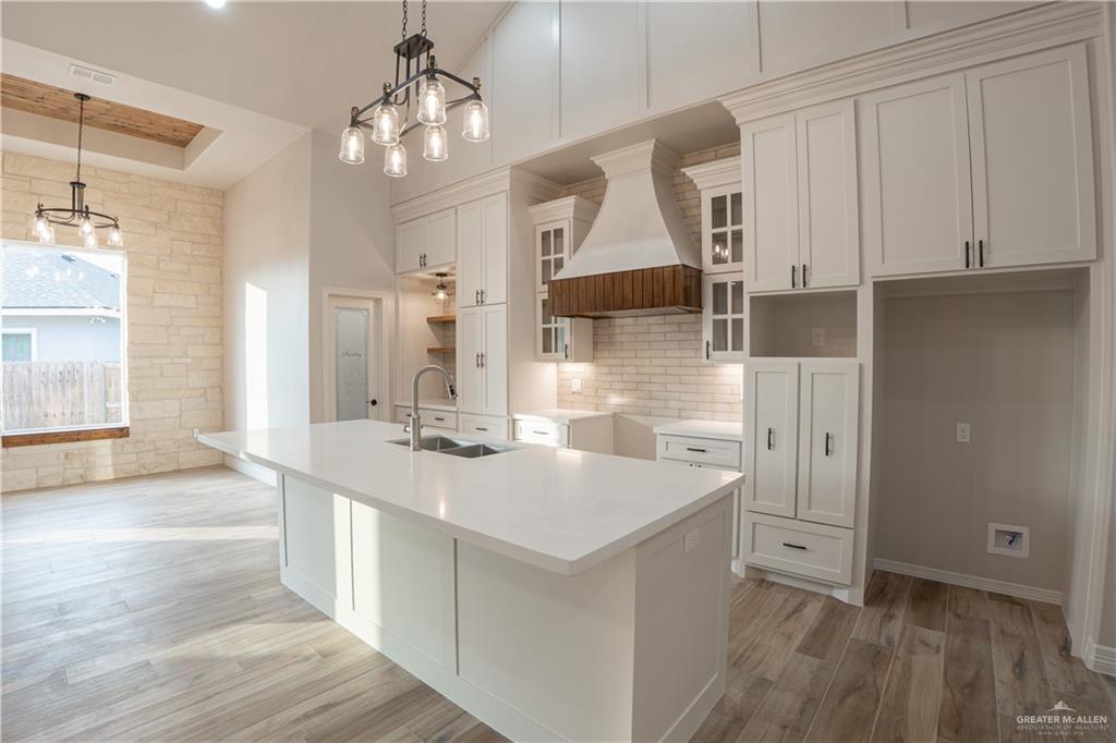 2618 Max Street Harlingen, TX 78552 - Photo 9 of 35 Modern kitchen featuring a large central island with an undermount sink, white cabinetry with dark hardware, a wood-finish range hood, and light brick-style backsplash
