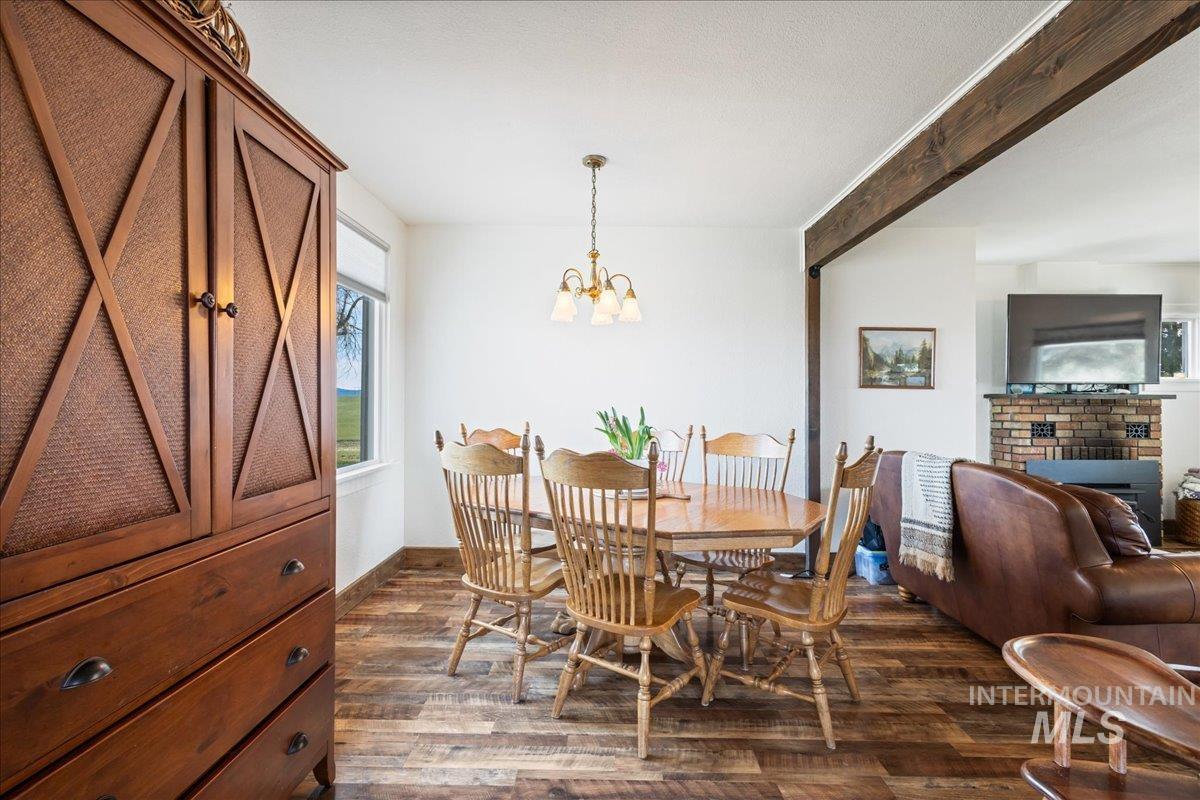 19454 Idaho Road Worley, ID 83876 - Photo 11 of 39 Dining area featuring wood-finish flooring, a brass-toned chandelier, a window offering exterior views, an exposed wood beam, and a partial view of a brick-clad fireplace