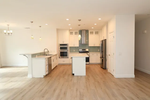 an empty room with wooden floor fireplace and kitchen view