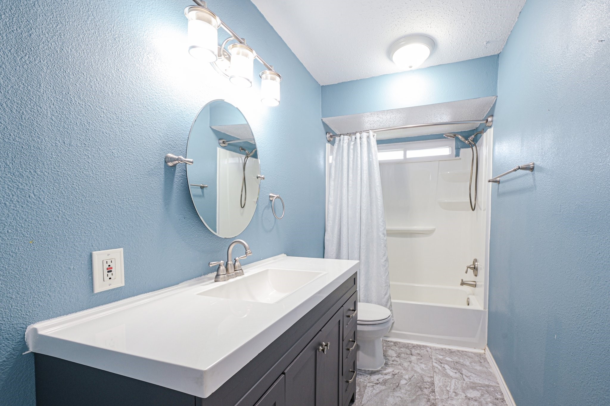 8 Arlington Street Conroe, TX 77301 - Photo 27 of 37 This bathroom features a modern design with blue walls, a sleek white countertop, a round mirror, and a combination bathtub and shower. The space is well-lit with stylish fixtures and has gray tile flooring.