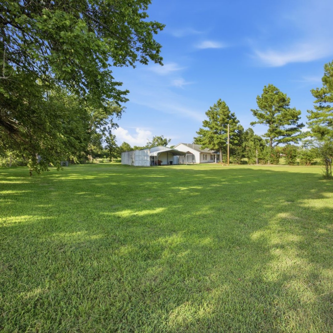 2696 Russellville Road Quinton, OK 74561 - Photo 39 of 40 Expansive green lawn with dappled sunlight, featuring mature trees and a distant residence with a prominent gabled roofline