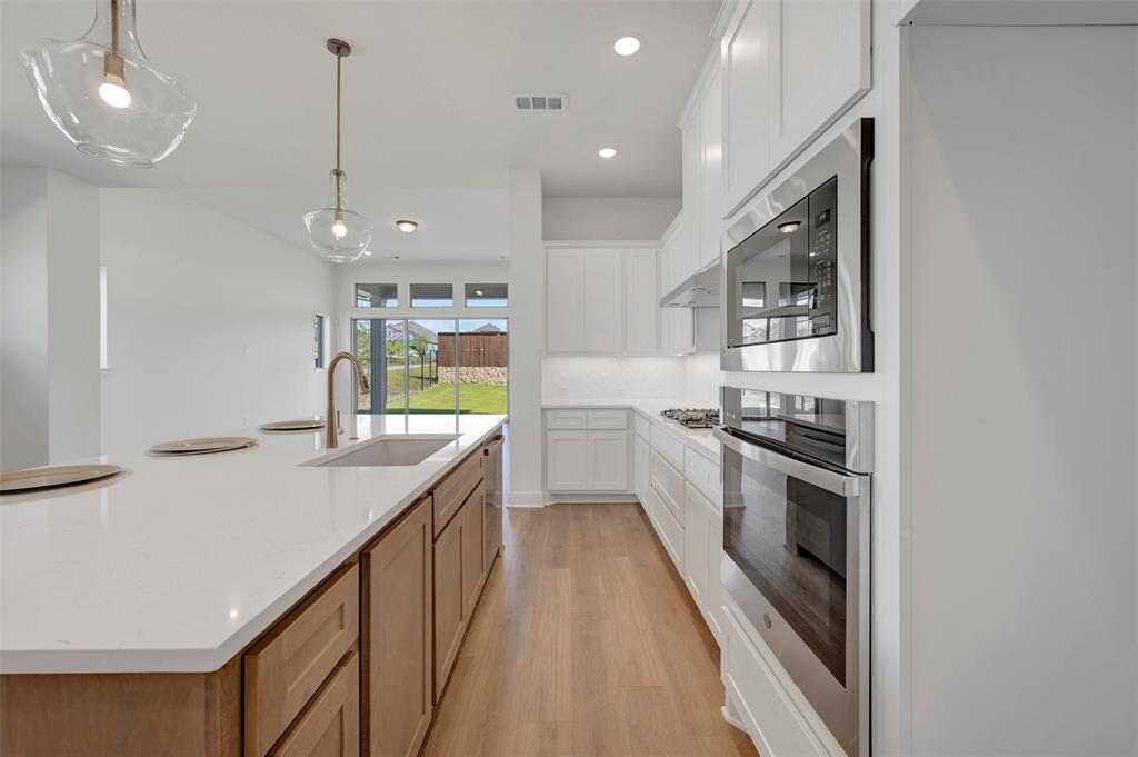 1351 Potenza Road Fate, TX 75087 - Photo 11 of 25 Open-concept kitchen featuring an island with a built-in sink and gold-finish faucet, light wood-finish flooring, white upper and lower cabinetry, stainless steel built-in microwave and oven, and recessed lighting
