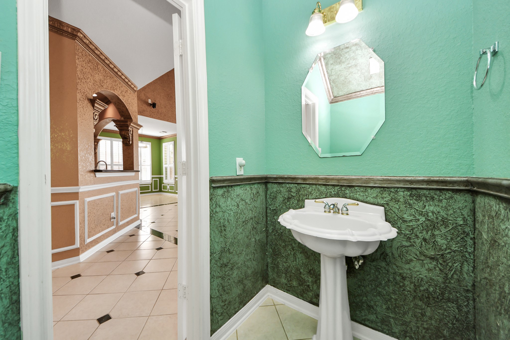 20111 Biscayne Hill Court Spring, TX 77379 - Photo 19 of 44 This photo features a small, elegantly decorated bathroom with a pedestal sink and octagonal mirror. The room is painted in a vibrant mint green with decorative molding. The view into the adjoining room reveals warm-toned walls, white trim, and tile flooring, offering a glimpse of the home's unique interior design.