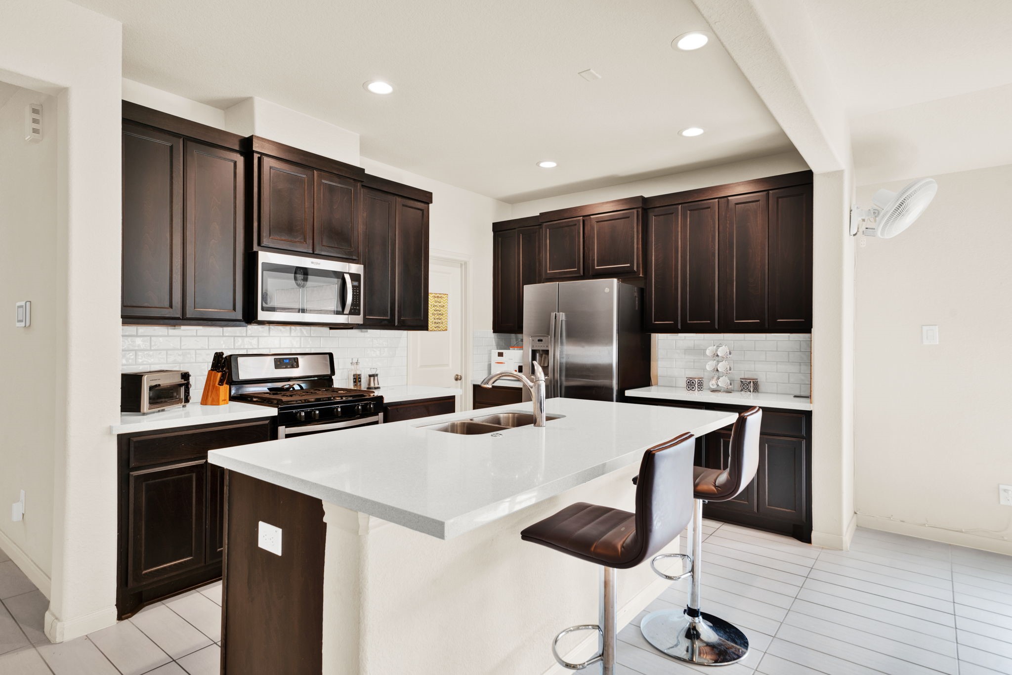 2503 Hagerman Road Conroe, TX 77384 - Photo 9 of 43 This kitchen features dark wood cabinets, a sleek white island with a built-in sink, and modern stainless steel appliances. The space is well-lit with recessed lighting and includes a tile backsplash and two bar stools for casual dining.