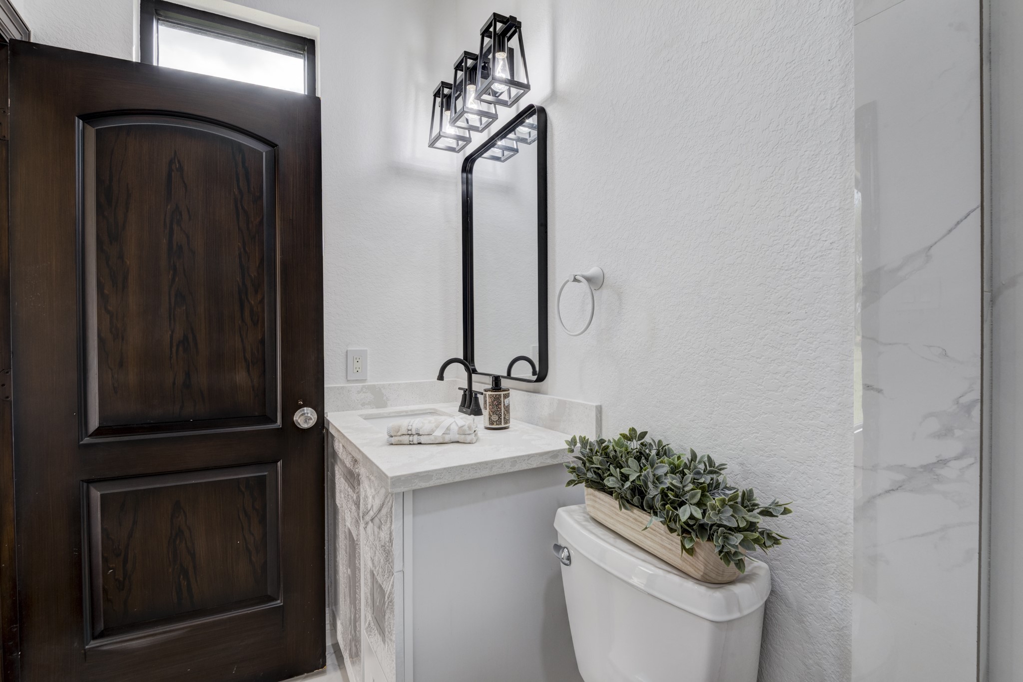 7348 Love Road Fulshear, TX 77441 - Photo 22 of 48 Elegant bathroom with a dark wood door, modern light fixtures, and a sleek vanity featuring a large mirror. The light walls and marble accents create a bright, sophisticated atmosphere.