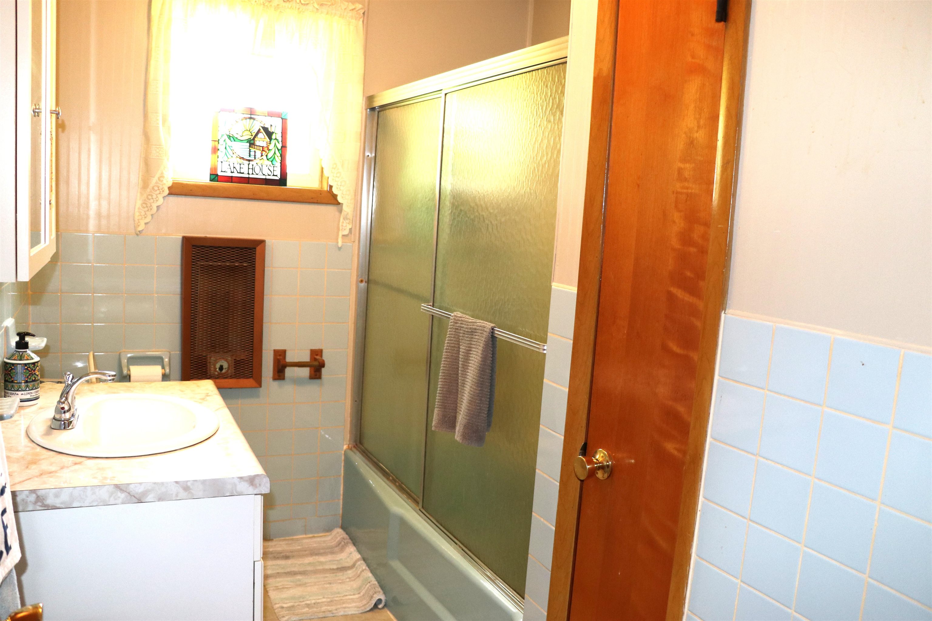 160 Patrick Drive Springville, TN 38256 - Photo 12 of 34 Bathroom featuring a vanity with an integrated sink, a full-size bathtub with a sliding glass enclosure, light blue ceramic wall tiling, a built-in recessed wooden cabinet, and a window with a valance