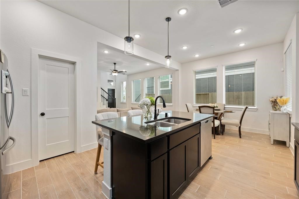 10014 Nectar Path Iowa Colony, TX 77583 - Photo 10 of 26 This photo showcases a modern, open-concept kitchen and dining area with sleek lighting. The kitchen features a dark island with a sink, complemented by light wood flooring. Large windows provide ample natural light, and there's a cozy living space visible in the background.