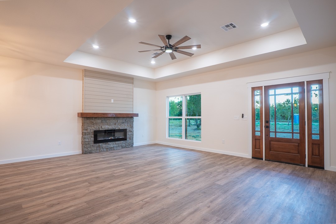 205 Blessings Way Colbert, OK 74733 - Photo 8 of 24 Wood-finish flooring, a stone-clad fireplace with a wood mantel, a multi-paneled wood entry door with glass, a tray ceiling, and recessed lighting