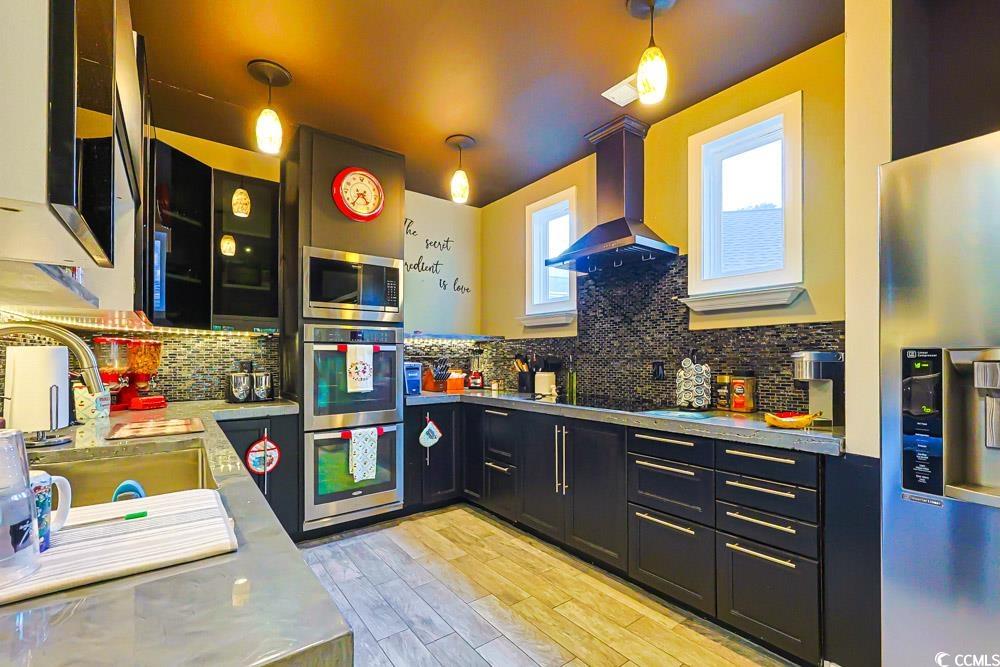 105 Rocko Drive Myrtle Beach, SC 29579 - Photo 4 of 38 Kitchen with dark cabinets, stainless steel appliances, wall chimney range hood, light wood-style floors, and tasteful backsplash