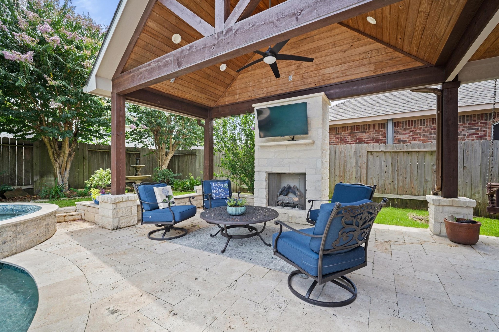 9507 Empress Crossing Drive Spring, TX 77379 - Photo 43 of 50 This inviting outdoor space features a covered patio with a wooden ceiling and fan, a cozy stone fireplace with a mounted TV, and comfortable seating. The area is beautifully landscaped, offering privacy with lush greenery. Perfect for entertaining or relaxing by the pool.