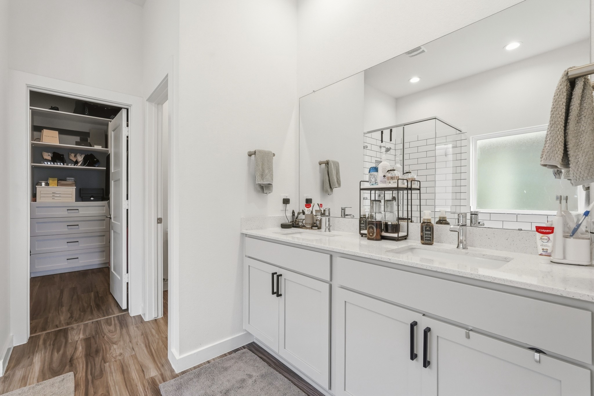 22522 Modica Rift Lane Katy, TX 77449 - Photo 22 of 38 The vanity side of the primary bath continues the clean, spa-inspired aesthetic. Crisp white shaker cabinets with matte black hardware support a wide quartz countertop with dual sinks — because great mornings are better shared. A full-width mirror reflects the light and space beautifully, and the walk-in closet just beyond the doorway offers built-in shelving and drawer storage that makes getting organized feel genuinely effortless.