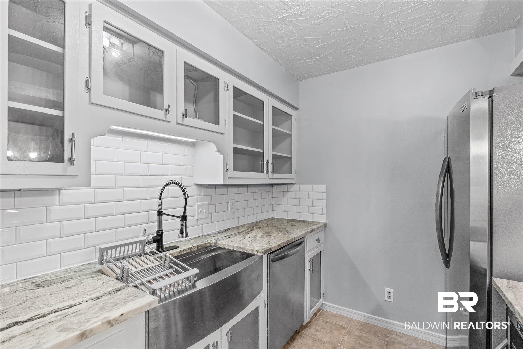 500 Grant Street, Unit C210 Daphne, AL 36526 - Photo 12 of 26 Kitchen featuring white shaker-style cabinetry with glass fronts, a stainless steel apron-front sink with a gooseneck faucet, light-toned granite countertops, a white subway tile backsplash, and stainless steel appliances
