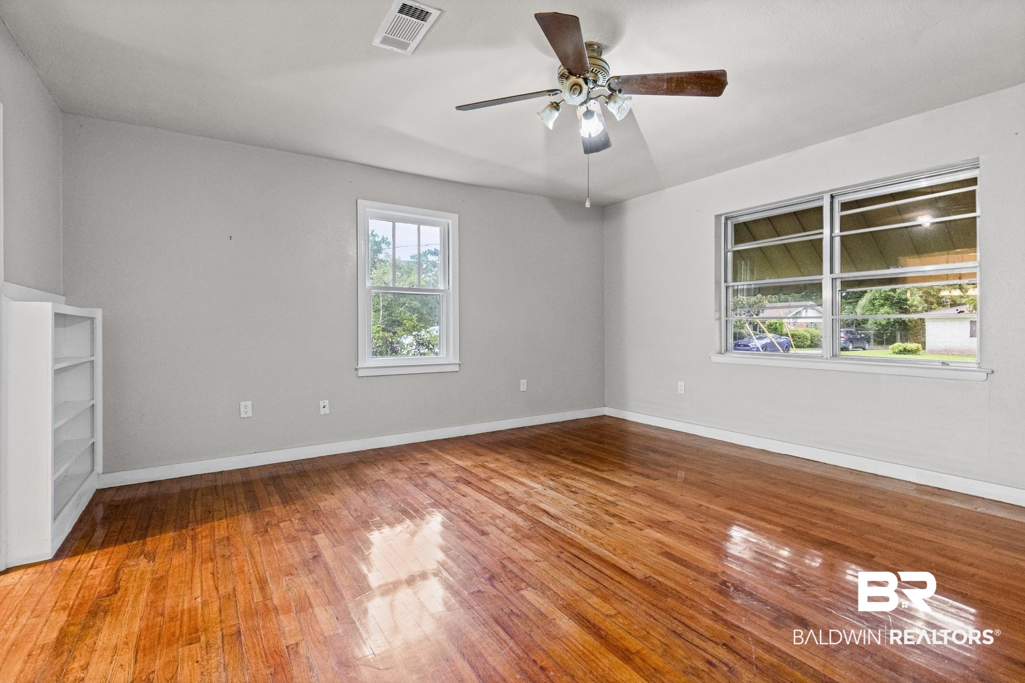 312 Sutherland Drive Mobile, AL 36611 - Photo 10 of 28 Spacious and unfurnished living room featuring wood-finish flooring, two windows providing natural illumination, a ceiling fan with integrated lighting, and a built-in white shelving unit