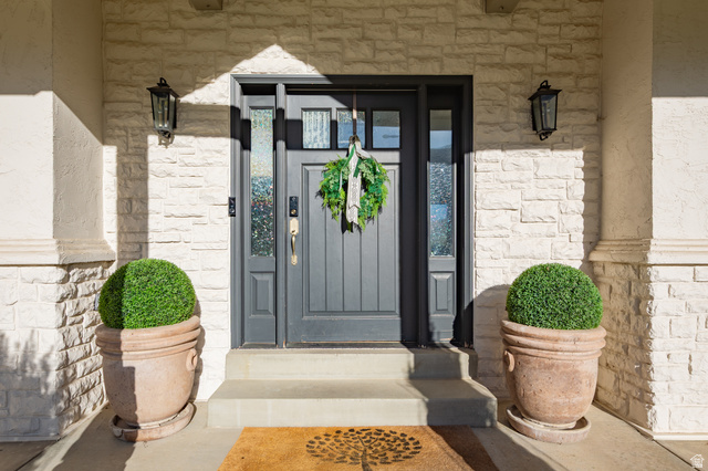 868 Braddock Lane Alpine, UT 84004 - Photo 6 of 64 Entryway featuring a dark gray paneled door with glass sidelights, textured stone veneer, stucco columns, concrete steps, and two black wall-mounted lanterns