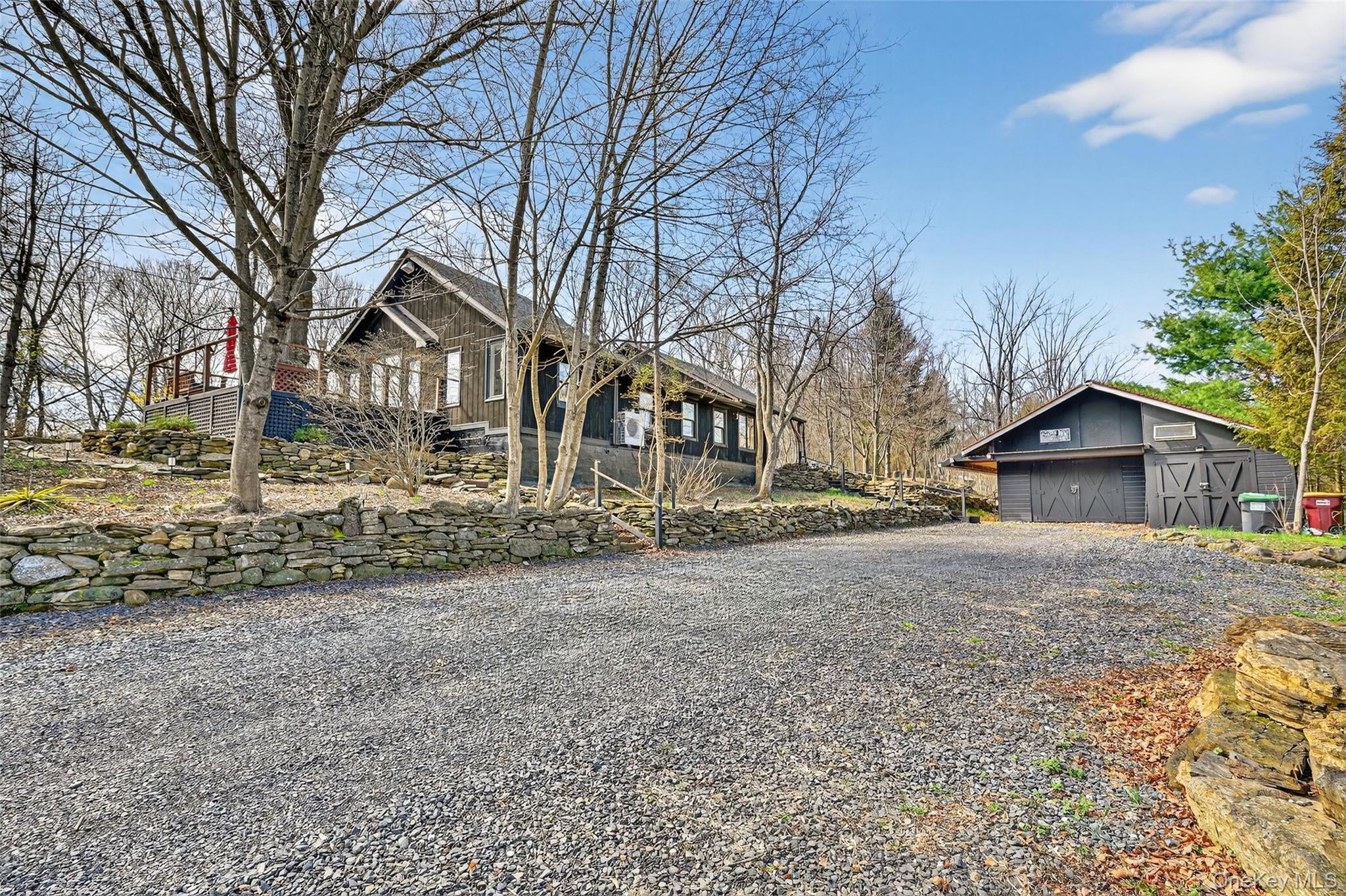 96 Lower Wisner Road Warwick, NY 10990 - Photo 2 of 34 Gravel driveway servicing a property featuring a dark-toned residence with an elevated deck, a stone retaining wall, and a separate dark-toned outbuilding with two garage doors