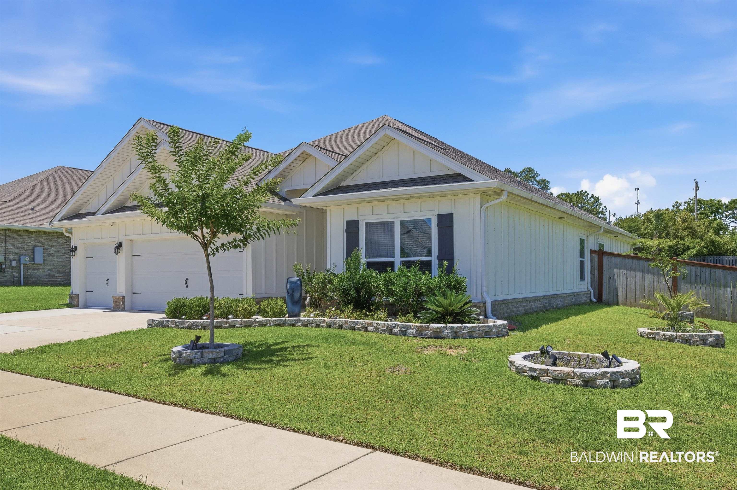 8735 Bronze Lane Foley, AL 36535 - Photo 47 of 58 White siding exterior with a two-car garage, mature landscaping, decorative window shutters, a front-facing window, and a stone-lined fire pit
