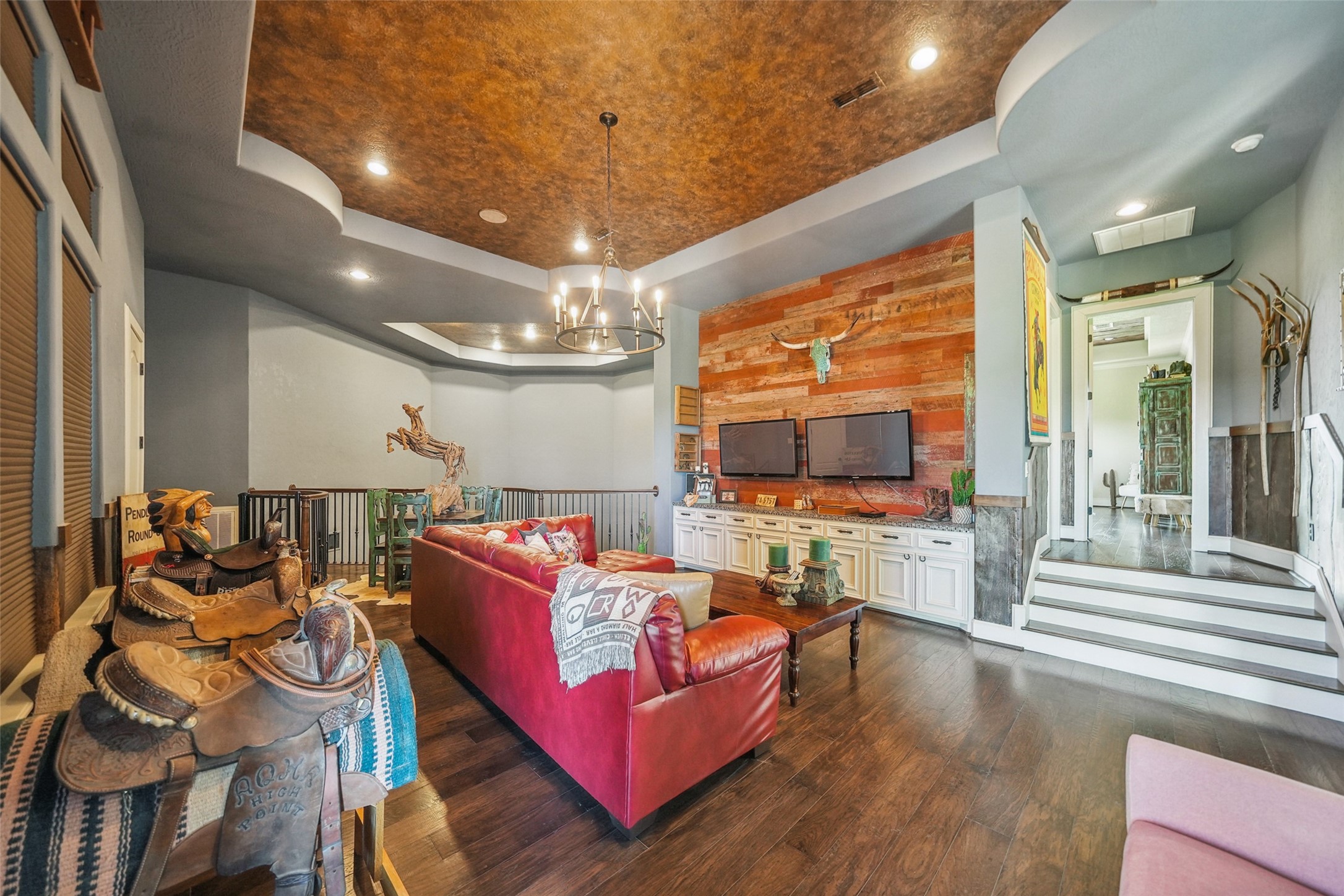 460 Bishop Road Huntsville, TX 77320 - Photo 24 of 50 This upstairs media lounge overlooks the theater room below and features reclaimed barnwood accents, faux-painted ceilings, and built-in cabinetry. Dual wall-mounted TVs create a perfect gaming setup, while a balcony showcases scenic ranch views. Pressed tin details add charm, and remote-controlled blackout blinds enhance the viewing experience. A dedicated media closet with built-in shelving and ventilation houses the home's AV equipment.