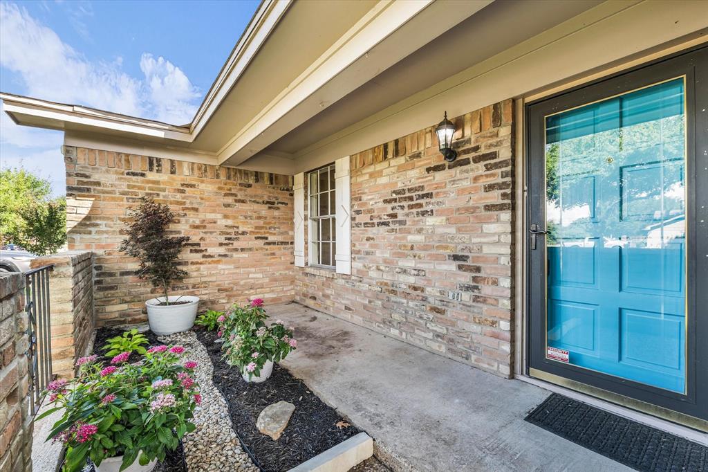 141 Travis Lane Hewitt, TX 76643 - Photo 3 of 31 Brick exterior featuring a vibrant blue entry door with a storm door, an outdoor wall sconce, a window with white shutters, and a landscaped garden bed with decorative rock and flowering plants