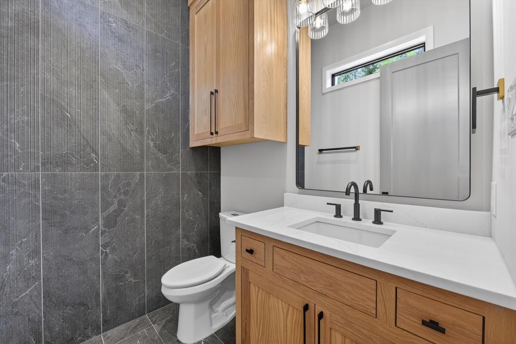 11028 Edgemere Road Dallas, TX 75230 - Photo 22 of 40 Powder Bathroom featuring dark grey tiled walls and flooring, a light wood-finish vanity with a white countertop and undermount sink, a large rectangular mirror, and powder off game/ living with overhead light fixture with glass shades