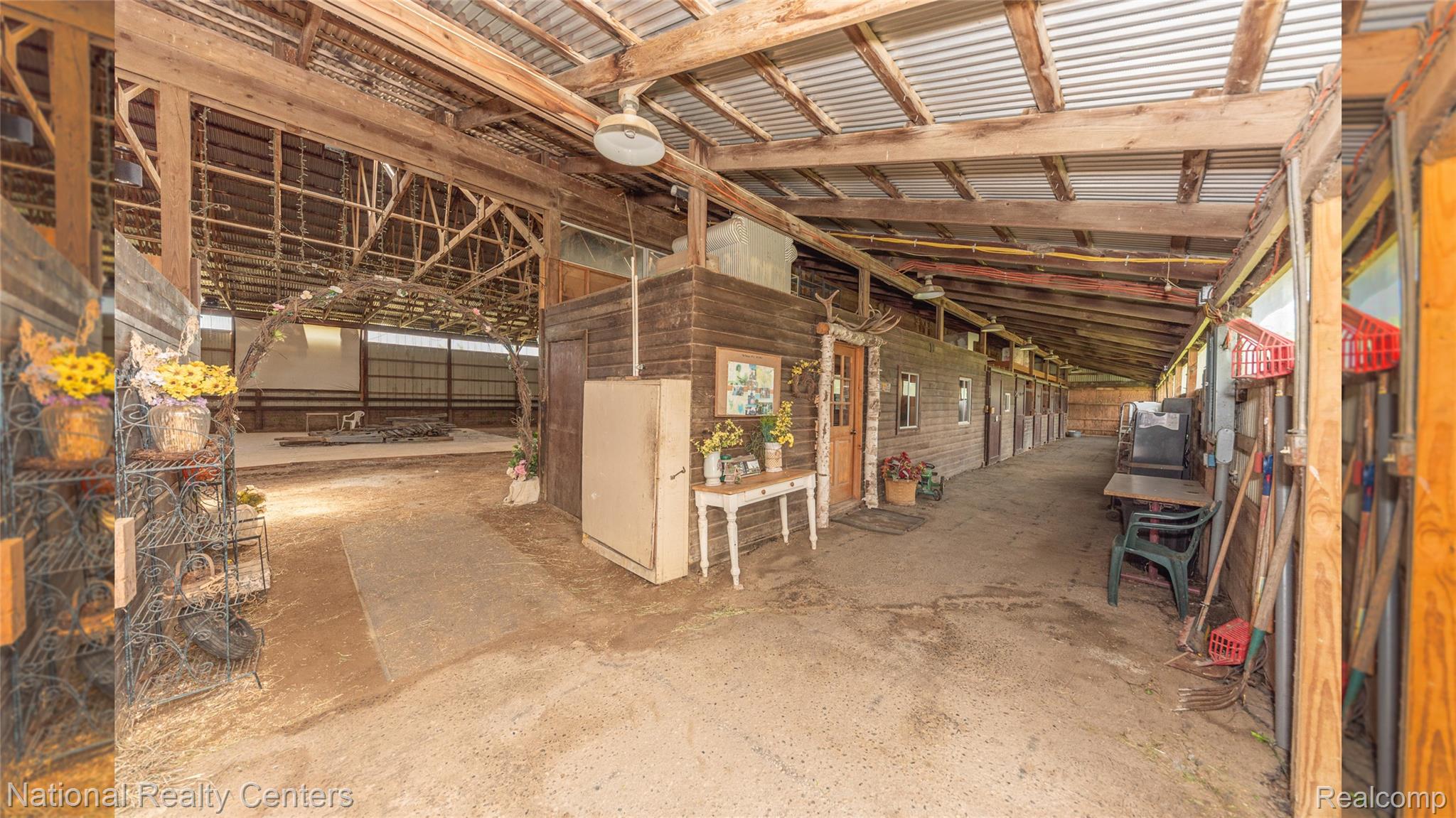 58191 8 Mile Road Northville, MI 48167 - Photo 22 of 57 Barn interior featuring a corrugated metal roof, exposed wood beam construction, multiple stalls with wood doors, and an open-air riding arena beyond